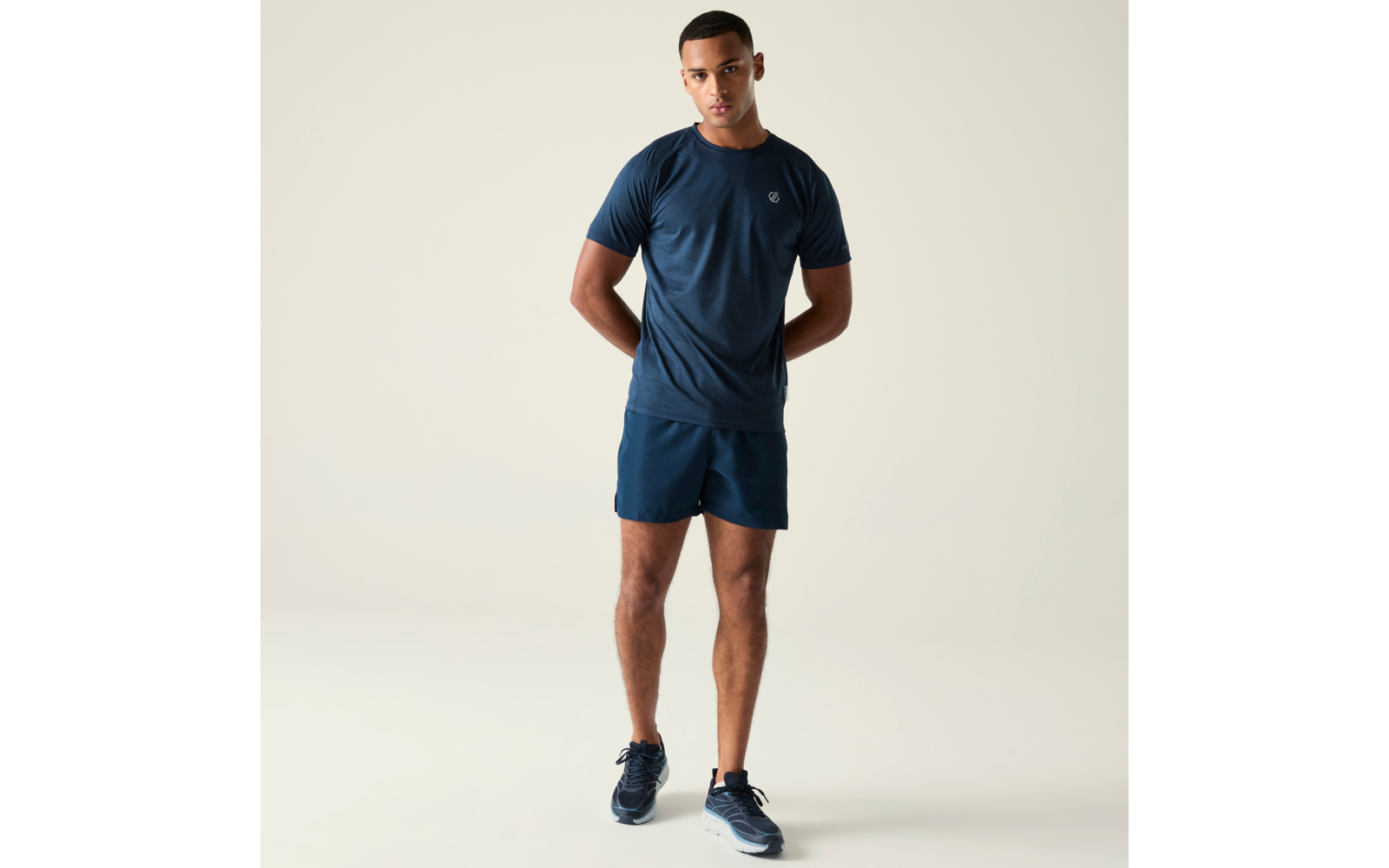 Homme debout en t‑shirt bleu foncé, short bleu foncé et baskets sur fond clair, mains dans le dos