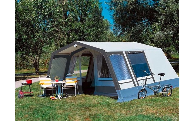Grote blauw-witte tent met open ingang, ervoor een kleine tafel met twee stoelen, een rode barbecue, een kleine fiets, gras, bomen en blauwe lucht