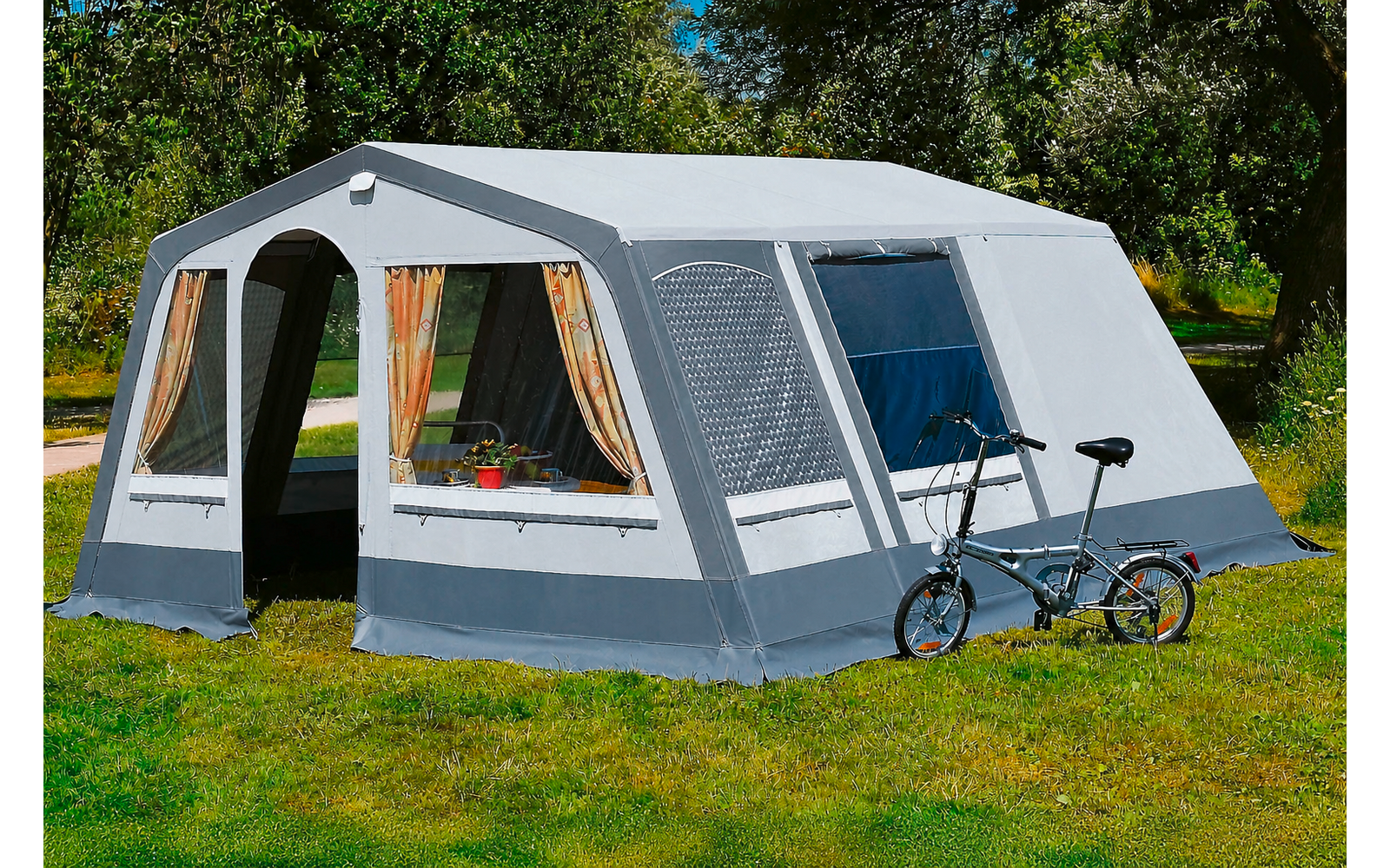 Grote grijs-witte tent op gras, twee ramen met gordijnen en bloempotten, open ingang, fiets leunt tegen de buitenkant, bomen op de achtergrond