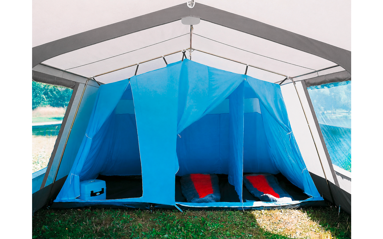 Binnenkant van een grote blauwe tent met open voorkant, drie slaapcabines, twee zichtbare rode slaapmatten en een blauwe koelbox op het gras