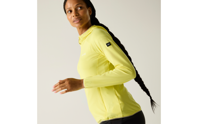 Seitliche Aufnahme einer Person mit langem geflochtenem Zopf, gelber Kapuzenjacke und schwarzer Leggings, die über die Schulter blickt vor hellem Hintergrund