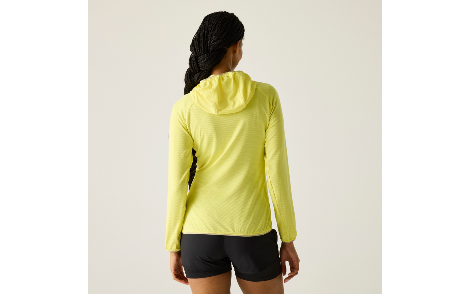 Person von hinten, trägt gelbe Kapuzenjacke und schwarze Shorts, geflochtene Haare, steht vor hellem Hintergrund