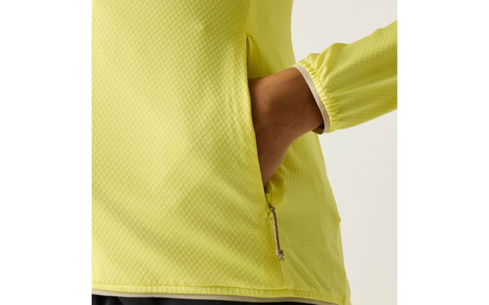 Nahaufnahme eines Oberkörpers in gelbgrünem, strukturiertem Langarmshirt; rechte Hand in einer seitlichen Reißverschlusstasche, sichtbarer Reißverschluss und Ärmelbündchen