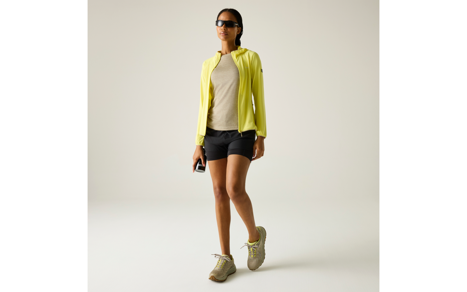 Person mit Sonnenbrille, gelber Jacke, beigem T‑Shirt, schwarzen Shorts und hellen Sportschuhen, hält ein Smartphone in der rechten Hand vor hellem Hintergrund