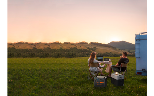 Ein Mann und eine Frau sitzen im Freien auf Stühlen vor einem Wohnmobil in einer grünen Wiese bei Sonnenuntergang