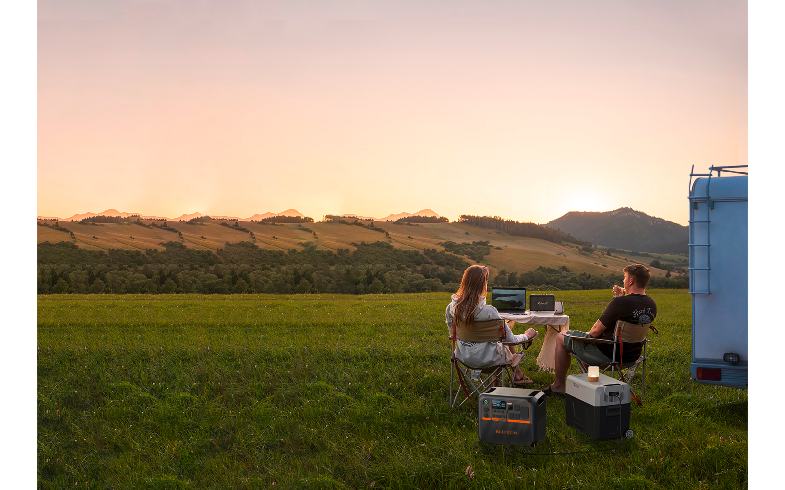 Ein Mann und eine Frau sitzen im Freien auf Stühlen vor einem Wohnmobil in einer grünen Wiese bei Sonnenuntergang