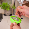 Twee handen houden een steelglas in een groene siliconenhouder op een aanrecht; op de achtergrond een gootsteen en een potplant