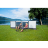 Zwei Personen sitzen auf Campingstühlen vor einem Windschutz an einem See, im Hintergrund Berge und ein Baum