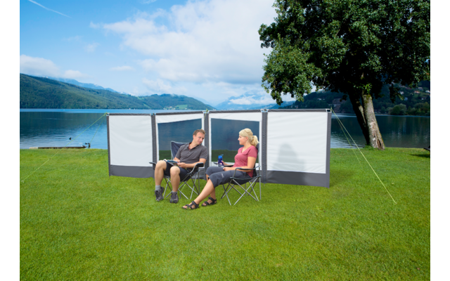 Zwei Personen sitzen auf Campingstühlen vor einem Windschutz an einem See, im Hintergrund Berge und ein Baum