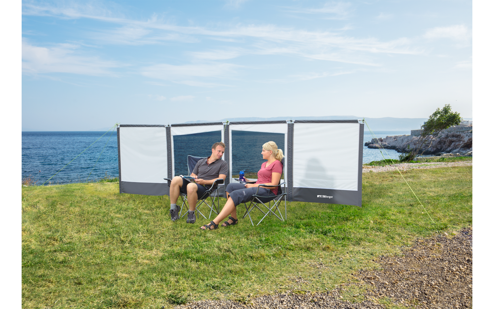 Zwei Personen sitzen auf Klappstühlen vor einer Windschutzwand auf einer Wiese am Meer