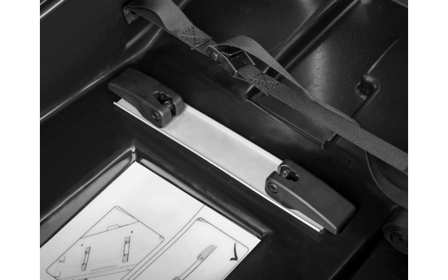 Innenansicht eines Kunststoffkoffers mit einer Metallschiene mit zwei schwarzen Kunststoffaufnahmen, einem schwarzen Gurt und einem Papierblatt mit schematischen Zeichnungen