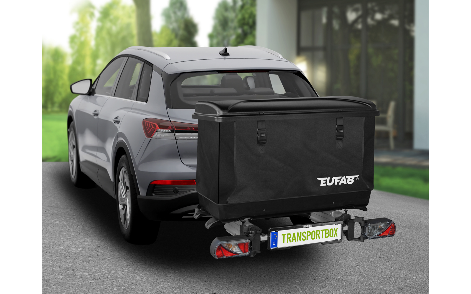 Grauer SUV von hinten links, an der Anhängerkupplung montierte schwarze Transportbox mit Aufdruck 'EUFAB' und Kennzeichen 'TRANSPORTBOX', Rückleuchten, Einfahrt, Haus mit Glasfront und Bäume im Hintergrund