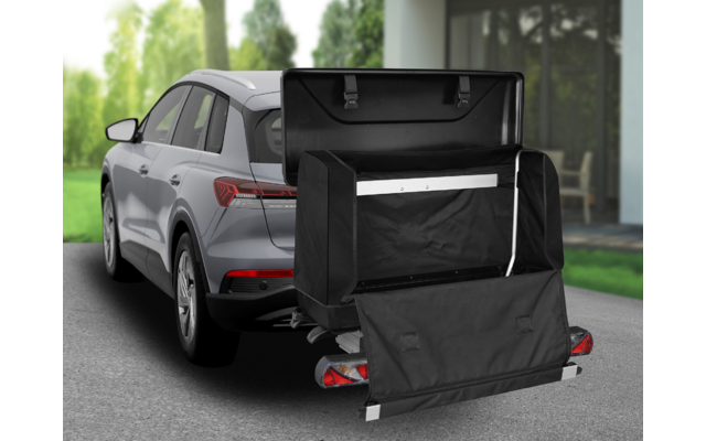 Grauer SUV von hinten links; an der Anhängerkupplung montierte schwarze, offene Gepäckbox mit heruntergeklappter Klappe; Metallträger; Haus und Bäume im Hintergrund
