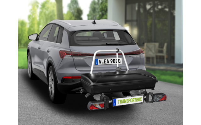 Grauer SUV mit am Anhängerkupplungs-Träger befestigter schwarzer Transportbox am Heck, Haus und Bäume im Hintergrund