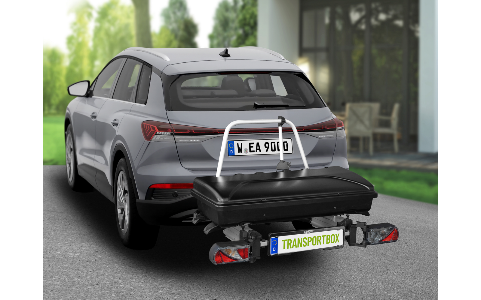 Grauer SUV mit am Anhängerkupplungs-Träger befestigter schwarzer Transportbox am Heck, Haus und Bäume im Hintergrund