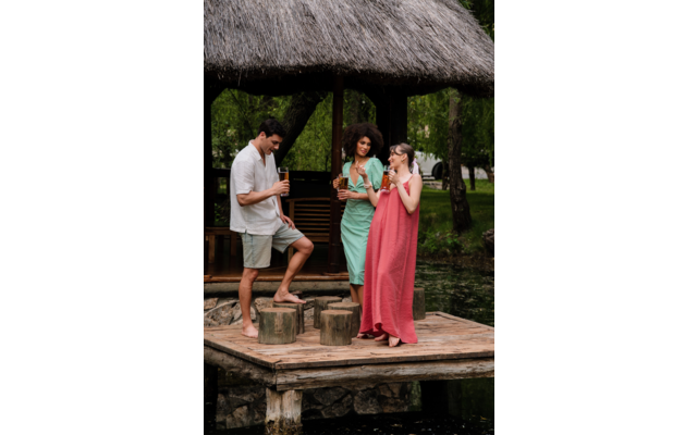 Trois personnes sur une jetée en bois sous un toit de chaume au bord de l'eau ; un homme est debout sur une souche, tous tiennent des boissons ; deux femmes portent de longues robes d'été (vert, rose)