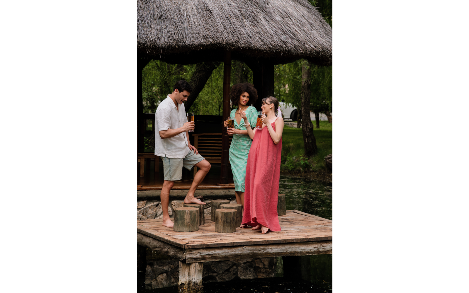 Trois personnes sur une jetée en bois sous un toit de chaume au bord de l'eau ; un homme est debout sur une souche, tous tiennent des boissons ; deux femmes portent de longues robes d'été (vert, rose)