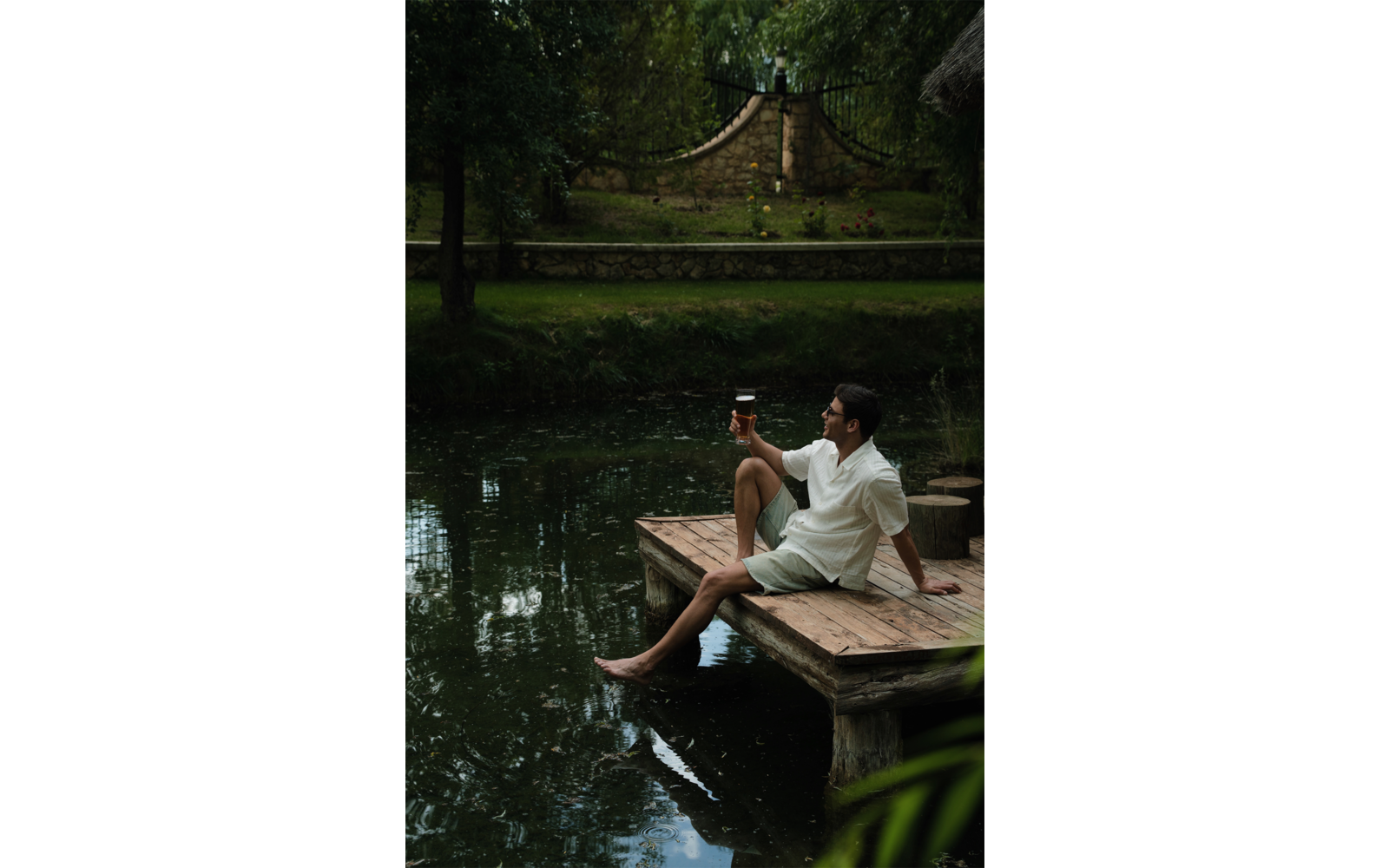 Personne en chemise blanche et short clair assise sur une jetée en bois au bord d'un étang, pieds dans l'eau, tenant un verre; en arrière-plan arbres, pelouse et un muret de pierre avec des marches