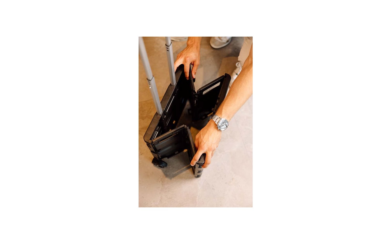 Deux mains tiennent une valise noire à roulettes ouverte avec la poignée télescopique sortie; montre-bracelet visible, chaussures blanches et sol clair