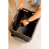 Deux mains sur une caisse noire pliable à roulettes sur un sol clair ; une poignée télescopique et des baskets grises visibles