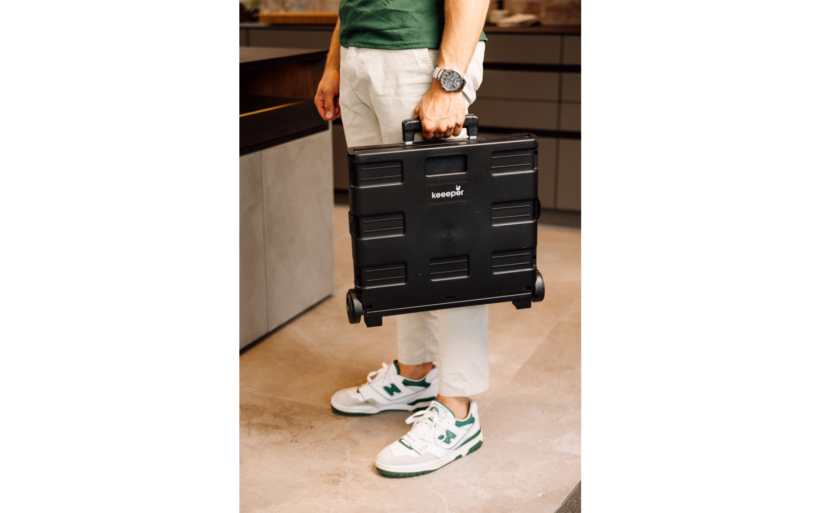 Partie inférieure d'une personne portant un haut vert, pantalon blanc et baskets blanches avec détails verts, tenant une mallette noire à roulettes, debout sur un sol carrelé clair à côté de placards de cuisine