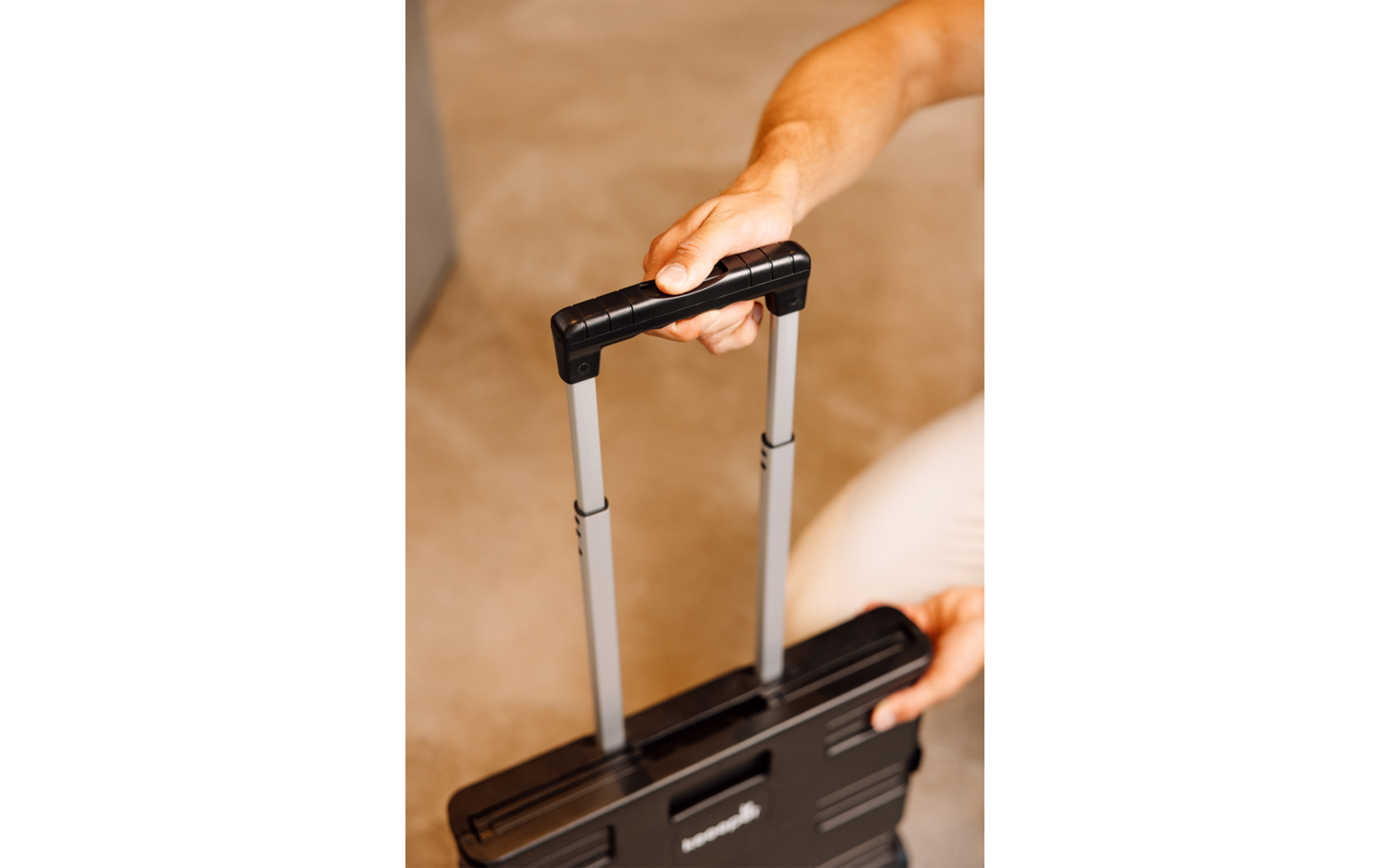 Mains tenant la poignée télescopique sortie d'une valise noire, sol beige en arrière-plan