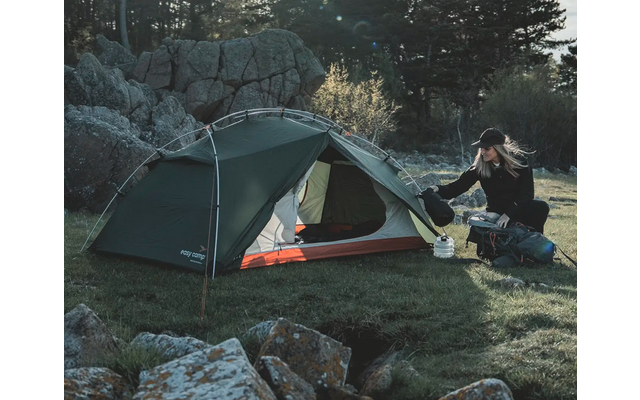 Zwei Personen beim Zelten in der Natur, neben einem grünen Zelt sitzt eine Person auf einer Decke