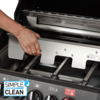Hand führt eine Edelstahlplatte mit Aussparungen in einen offenen Gasgrill ein; Brennerroste und Bedienfeld mit Drehknöpfen sichtbar, 'SIMPLE CLEAN'-Aufkleber unten links
