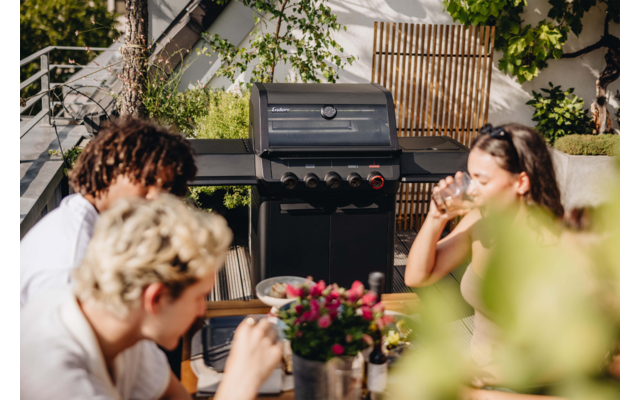 Vier Personen sitzen an einem Gartentisch und halten Getränke; im Hintergrund ein Gasgrill, Blumen auf dem Tisch