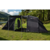 Zwarte tent met open ingang en groen interieur, luifel met stokken en scheerlijnen op gras aan een meer, bomen en heuvels op de achtergrond onder een bewolkte lucht