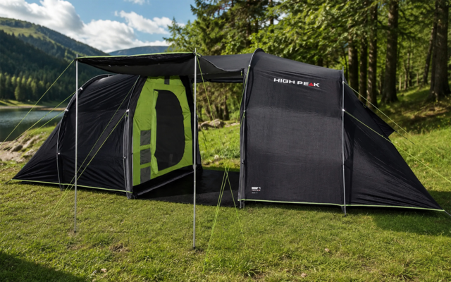 Zwarte tent met open ingang en groen interieur, luifel met stokken en scheerlijnen op gras aan een meer, bomen en heuvels op de achtergrond onder een bewolkte lucht