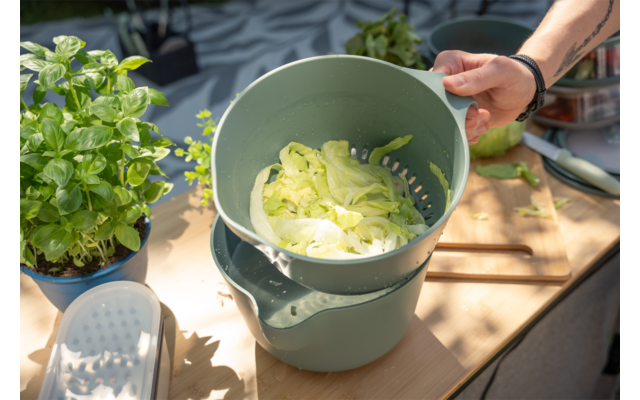 Rechterhand houdt een groene kunststof vergiet met dunne lichtgroene plakjes boven een kom; links een pot met basilicum, houten tafel