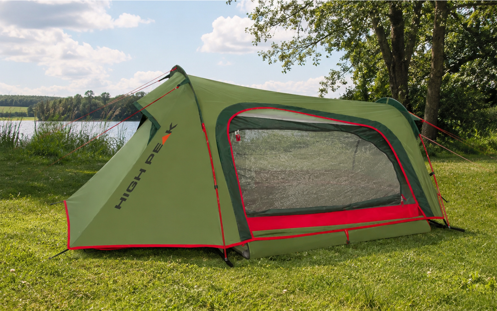 Green tent with red accents and a mesh side window on grass, lake and trees in the background under a partly cloudy sky