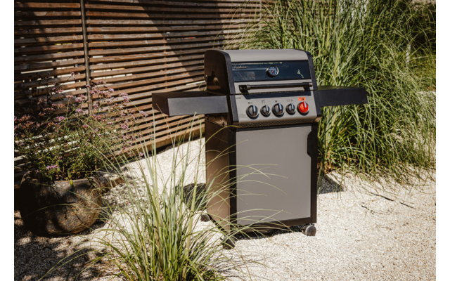 Gasgrill steht auf gepflastertem Boden vor einem Ziegelhaus, umgeben von Gras und Pflanzen