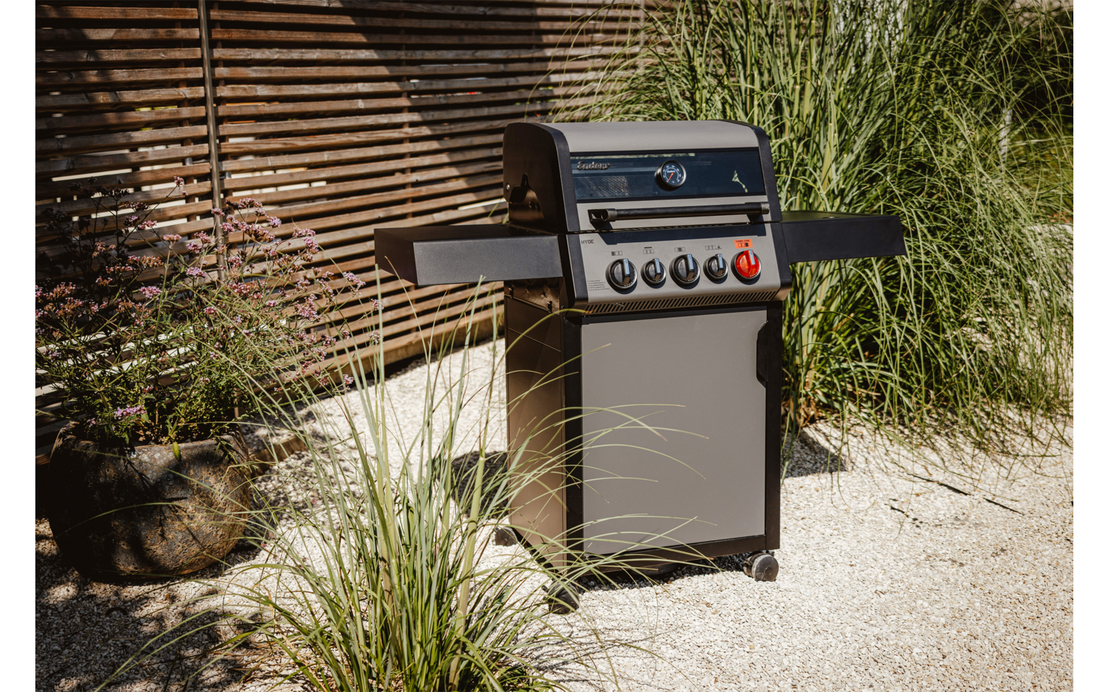 Gasgrill steht auf gepflastertem Boden vor einem Ziegelhaus, umgeben von Gras und Pflanzen