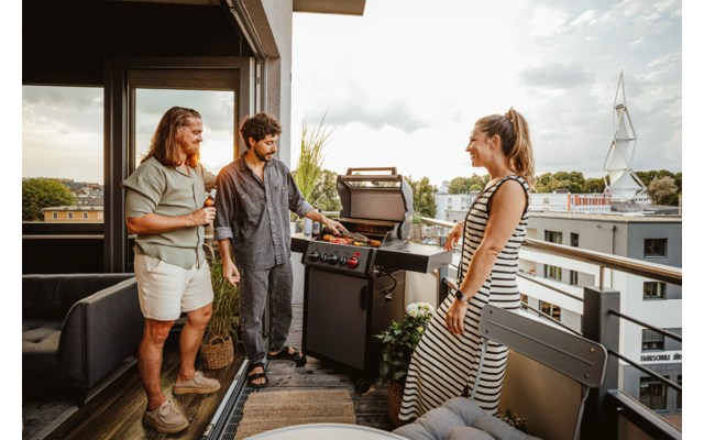 Drei Personen auf einem Balkon, eine Person bedient einen Grill