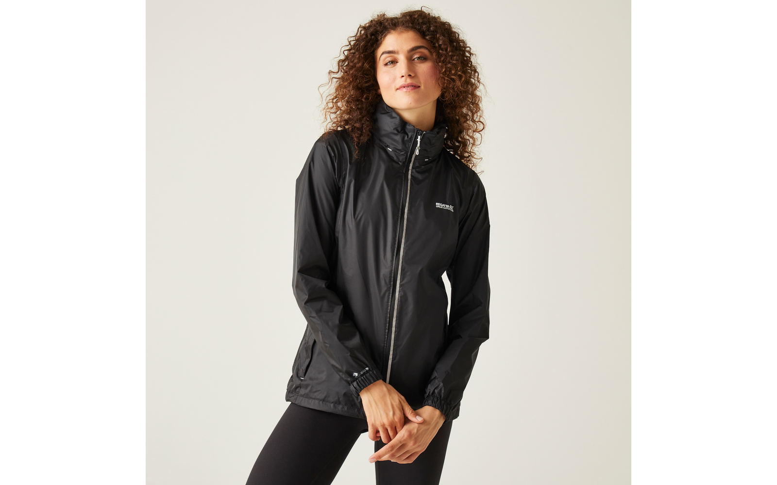 Persona con capelli ricci che indossa una giacca nera con cerniera e collo alto e piccolo logo bianco sul petto, mani davanti al corpo, sfondo chiaro uniforme