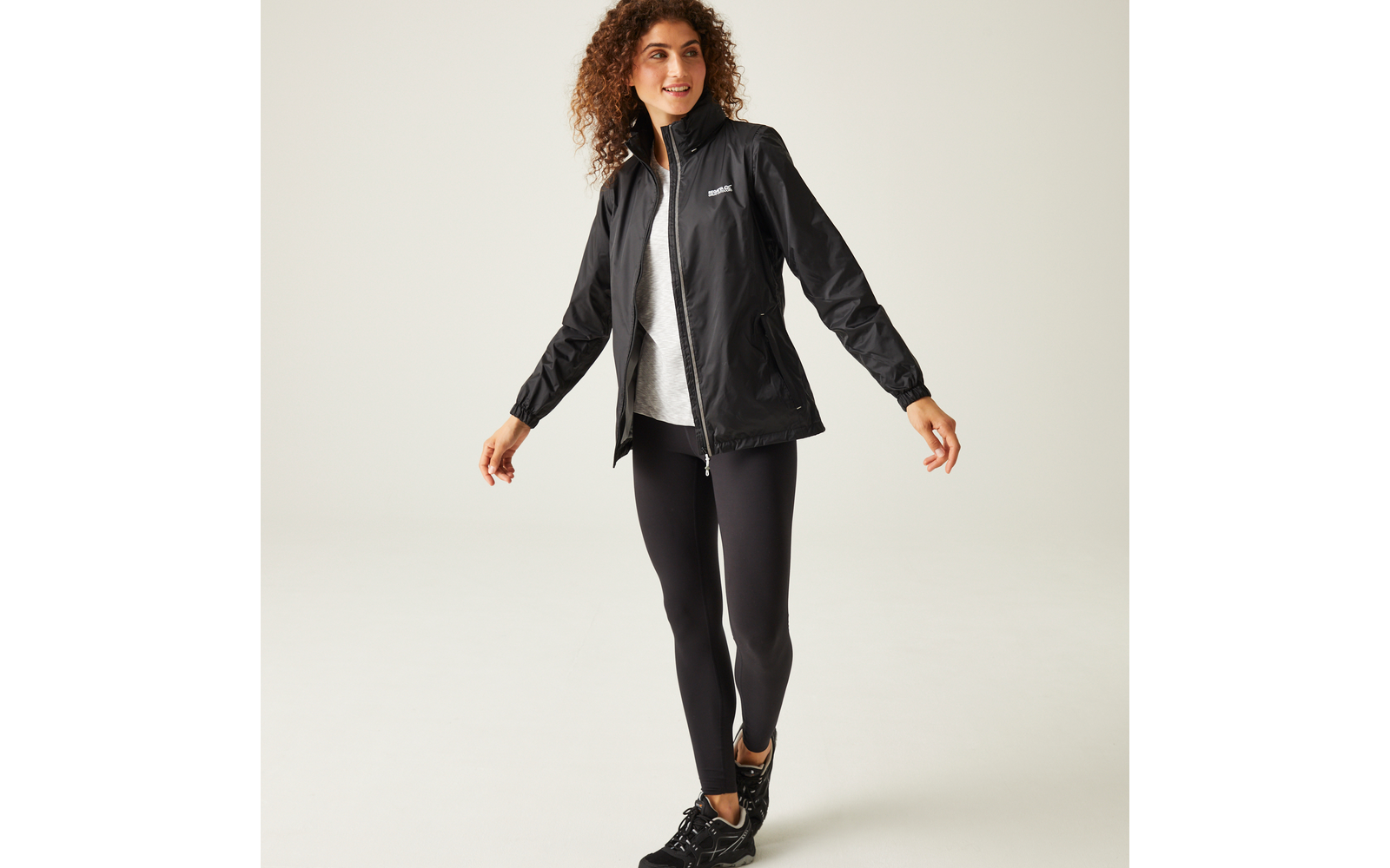 Persona con capelli ricci fino alle spalle che indossa una giacca nera, una maglia grigia, leggings neri e scarpe sportive nere, in piedi su uno sfondo chiaro uniforme