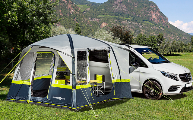 Campingwagen mit angebautem Zelt auf einer grünen Wiese, Berge und Bäume im Hintergrund