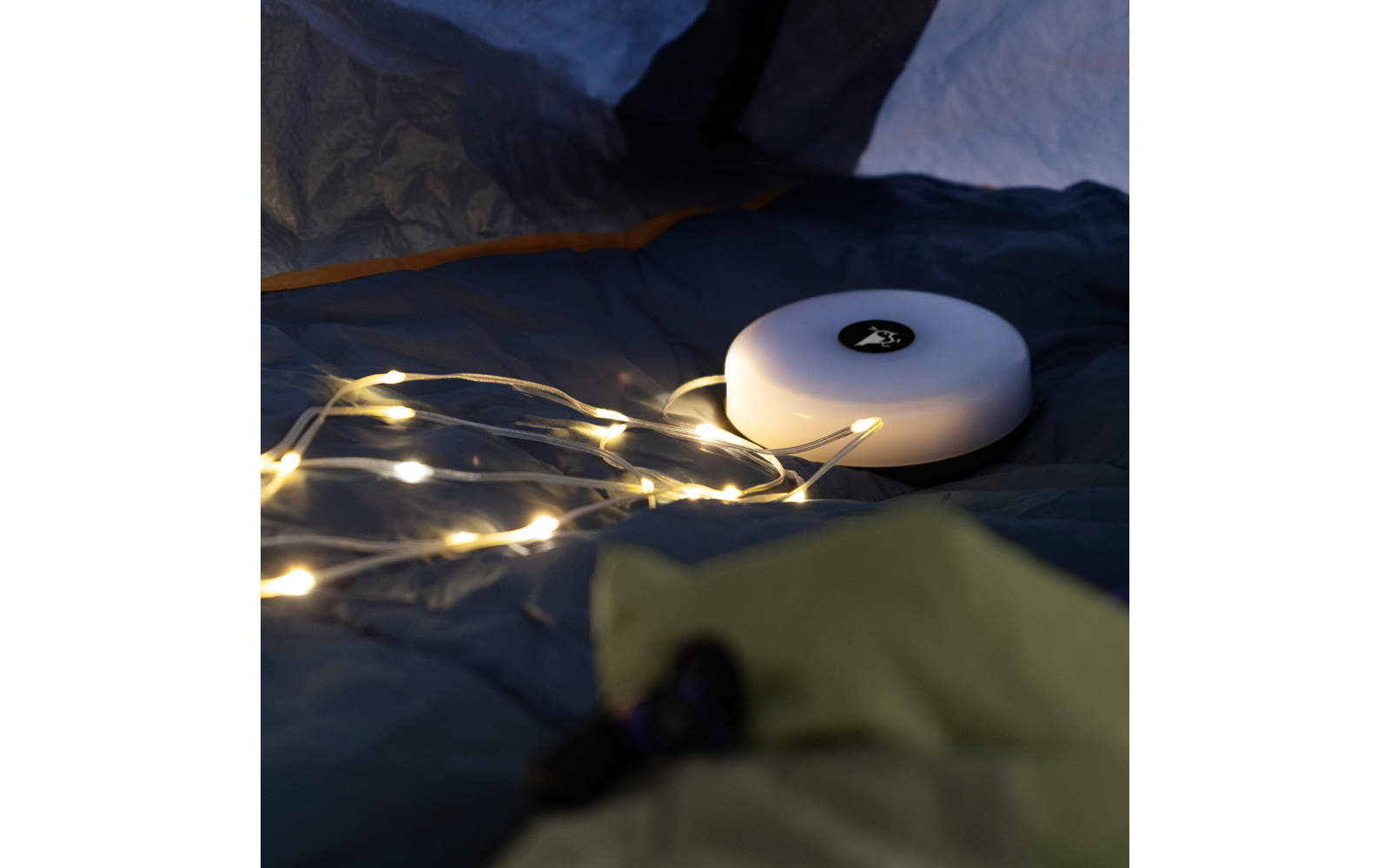 Rundes weißes Gerät mit schwarzem Symbol auf der Oberseite, liegend auf dunklem Stoff, daneben eine leuchtende Lichterkette mit warmweißen LEDs, im Vordergrund unscharfer grüner Stoff