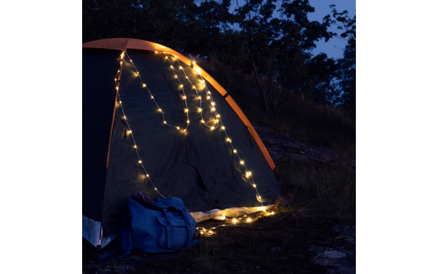 Zelt mit orangefarbener Kante, Lichterketten an der Vorderseite und ein blauer Rucksack davor; nächtlicher Himmel im Hintergrund
