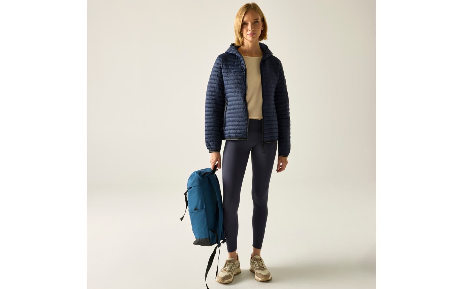 Person mit schulterlangem blondem Haar, trägt eine dunkelblaue gesteppte Jacke, helles Sweatshirt, dunkle Leggings und beige Sneaker, hält einen blauen Rucksack in der linken Hand, steht vor hellem Hintergrund