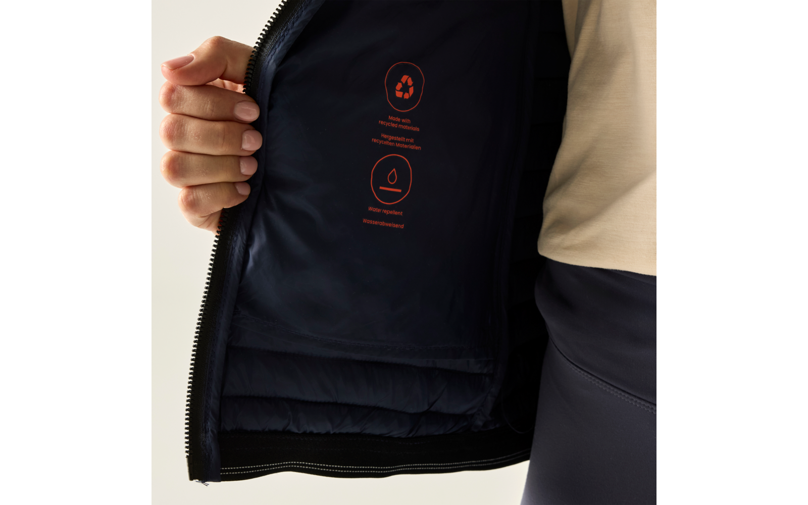 Hand hält eine geöffnete dunkelblaue Jacke mit offenem Reißverschluss; Innenfutter zeigt drei rote Symbole und kleine rote Textzeilen; sichtbarer Unterarm in beige Oberteil und dunkle Hose