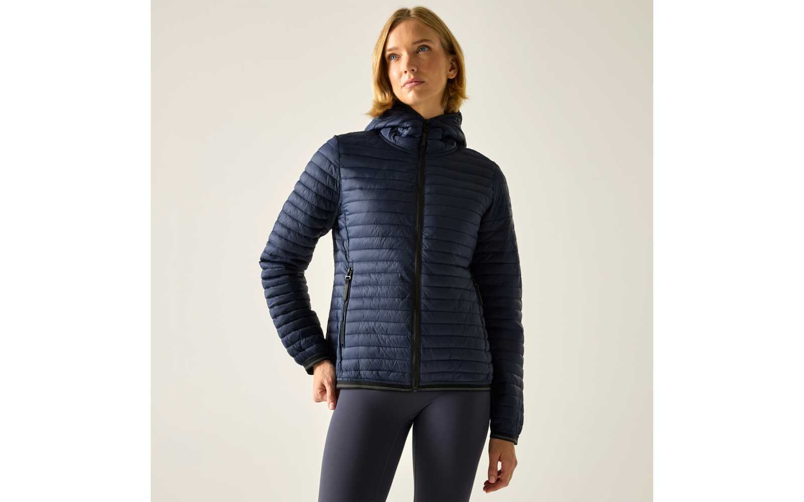 Person mit schulterlangen blonden Haaren trägt dunkelblaue gesteppte Kapuzenjacke mit Reißverschluss und dunkle Leggings, steht vor hellgrauem Hintergrund