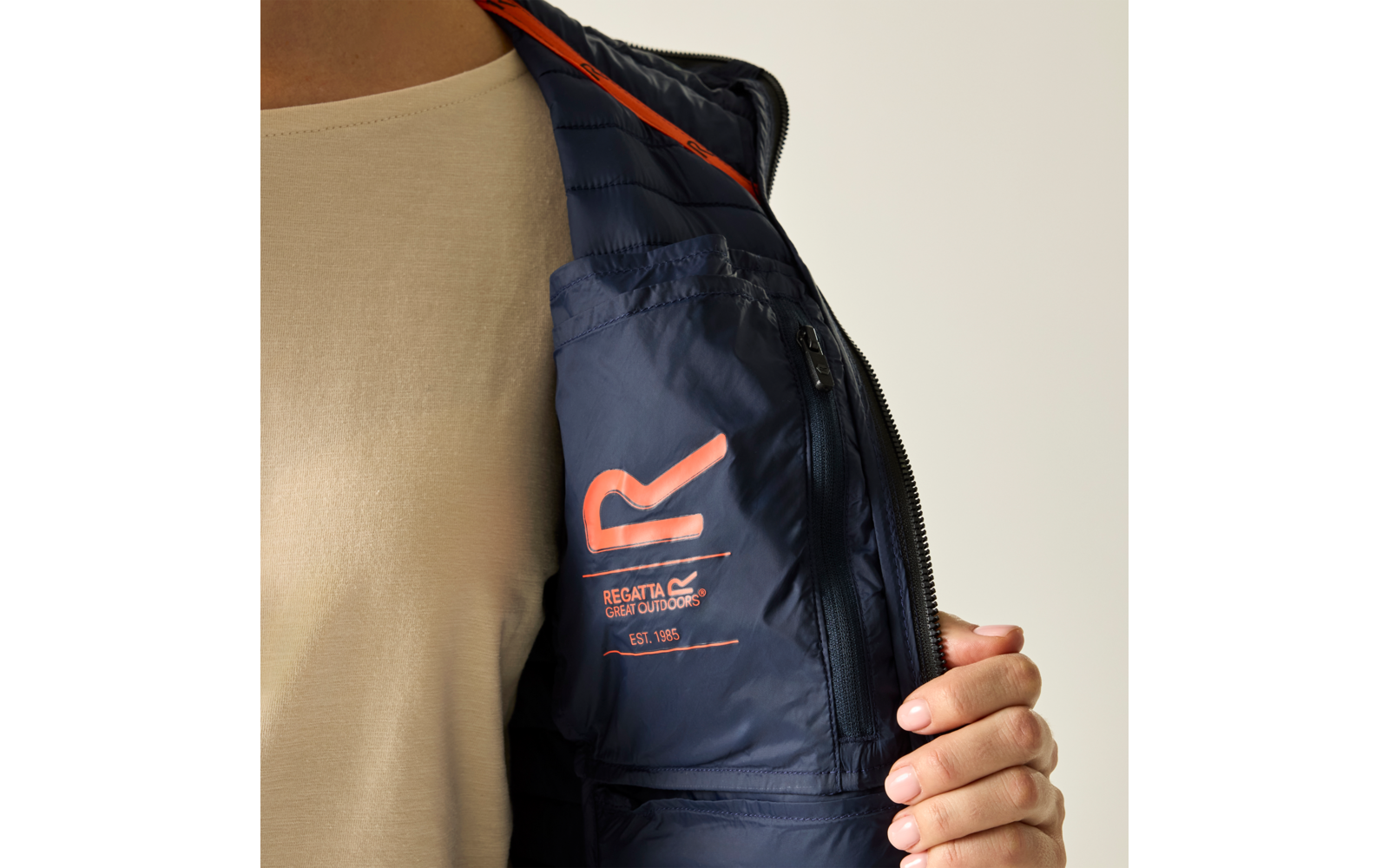 Oberkörper einer Person in beigem Shirt, hält eine geöffnete marineblaue Jacke mit sichtbarem orangefarbenem Logo und Reißverschluss