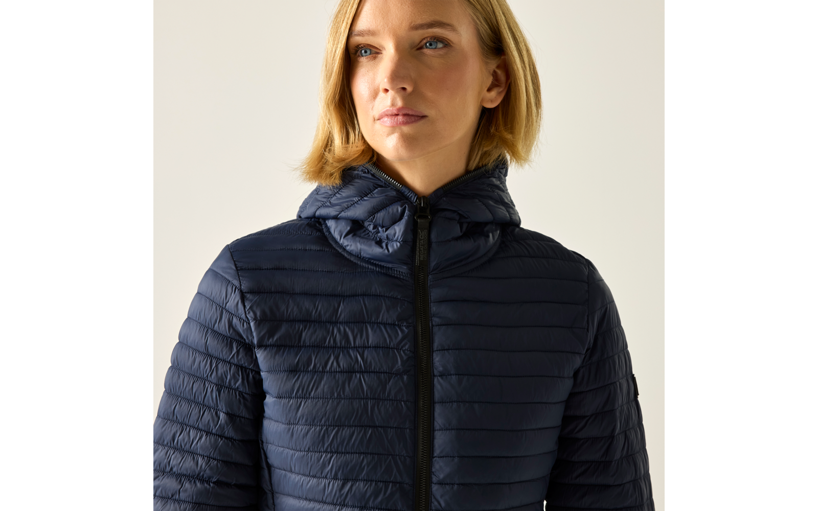 Person mit kinnlangen blonden Haaren trägt dunkelblaue gesteppte Jacke mit Kapuze und geschlossenem Reißverschluss, heller einfarbiger Hintergrund