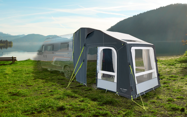 Wohnwagen steht auf einer grünen Wiese neben einem See, im Hintergrund Berge und blauer Himmel