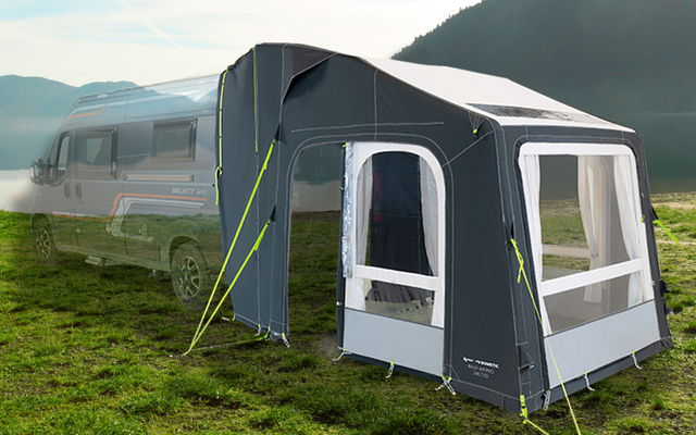 Wohnwagen steht auf einer grünen Wiese neben einem See, im Hintergrund Berge und blauer Himmel