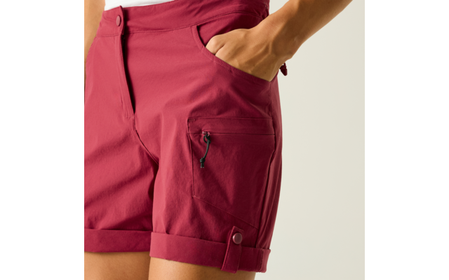 Burgunderrote Shorts, rechte Hand in seitlicher Eingrifftasche, sichtbare Reißverschlusstasche am Oberschenkel, umgeschlagener Saum mit Knopflasche, heller Hintergrund