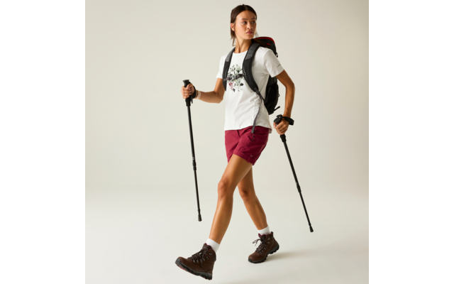 Person mit Zopf trägt weißes T‑Shirt mit Blumenmotiv, bordeauxrote Shorts, braune Wanderschuhe, weiße Socken, schwarzer Rucksack mit roter Innenfarbe; hält zwei schwarze Trekkingstöcke; heller einfarbiger Hintergrund